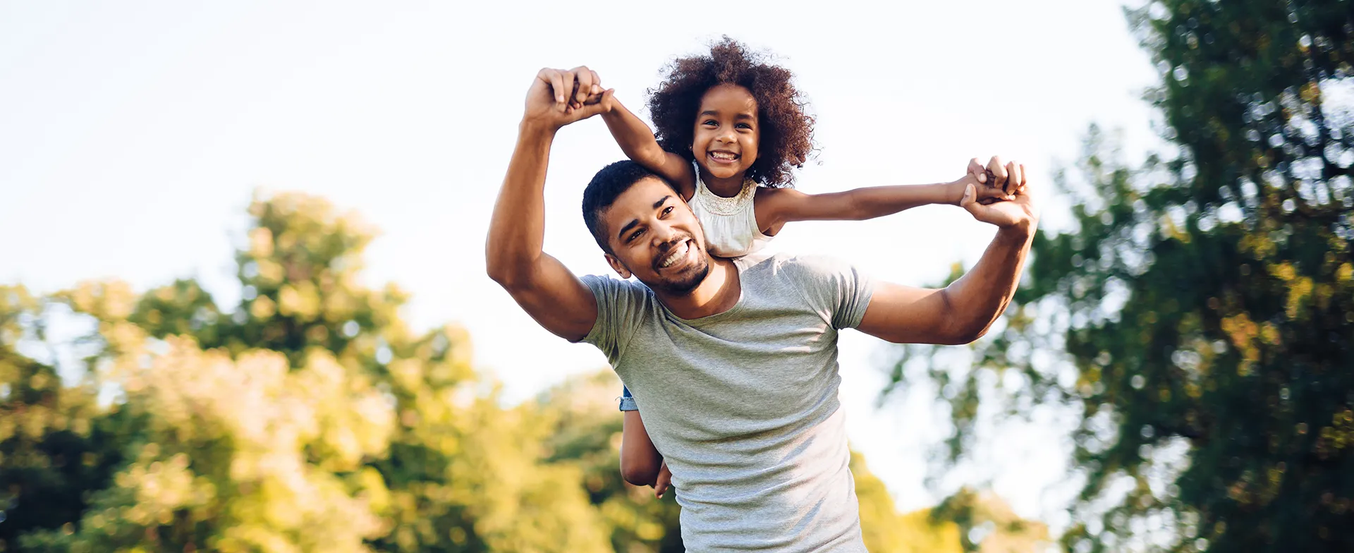A smiling father carrying his young daughter on his shoulders outdoors